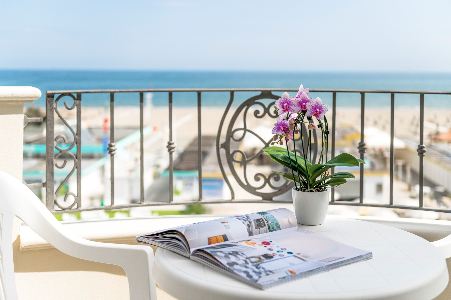 Balcon avec vue sur la plage, une orchidée en pot et un magazine sur une table blanche.