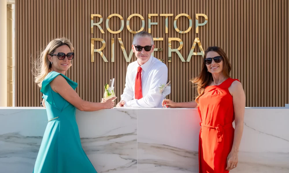 Drei Personen in eleganter Kleidung stehen vor einem Schild mit der Aufschrift "Rooftop Riviera" und genießen Cocktails.