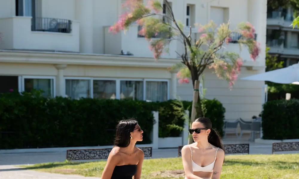 Zwei Frauen sitzen mit Einkaufstaschen vor einem Hotel auf einer Bank im Freien.