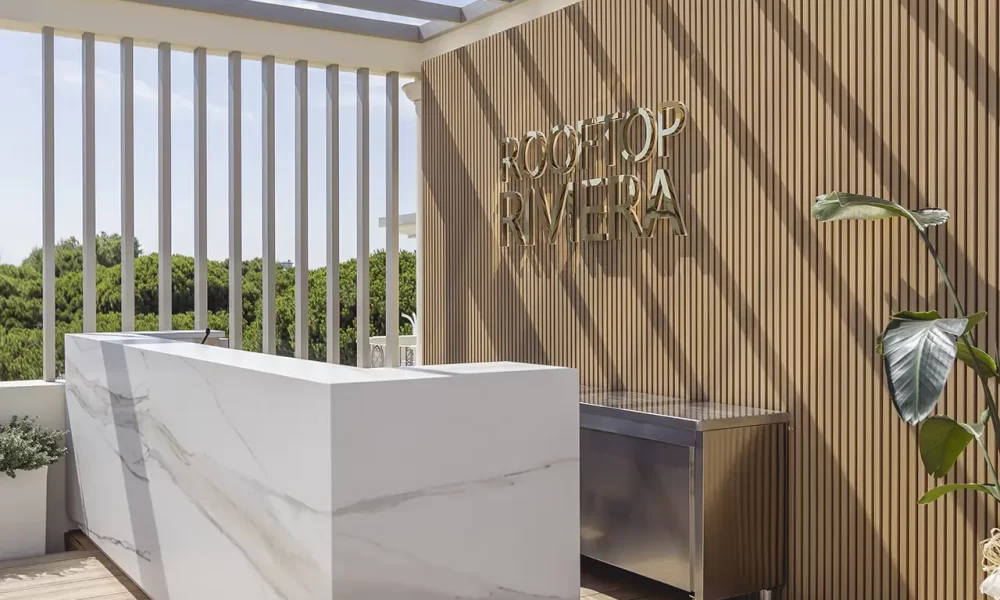 Terrasse moderne avec un bar en marbre et un panneau "Rooftop Riviera" sous une pergola.