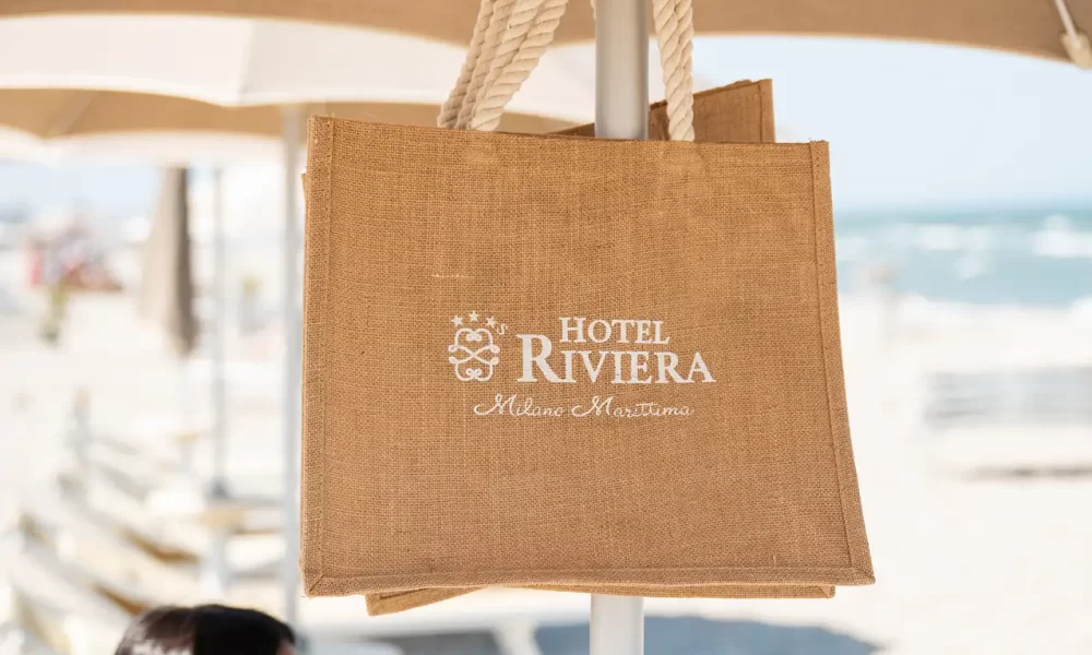 Eine Strandtasche vom Hotel Riviera hängt an einem Sonnenschirm, im Hintergrund ist eine Frau am Strand zu sehen.