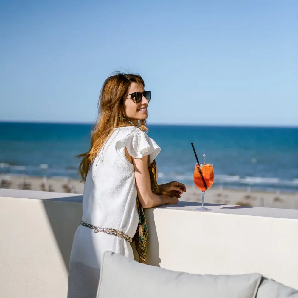 Frau am Strand genießt ein Getränk auf einer Terrasse mit Blick aufs Meer.
