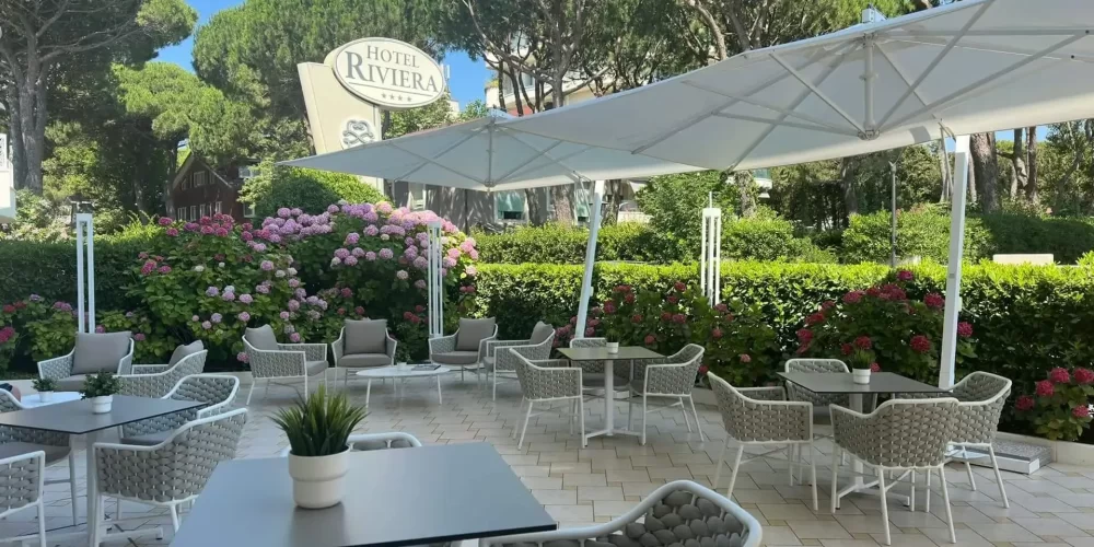 Terrasse extérieure de l'Hôtel Riviera avec des tables et des chaises entourées de fleurs et de verdure.